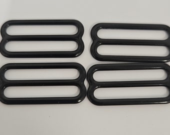 May include: Four black metal bra strap sliders, each with three loops. The sliders are arranged on a white surface. These are used for adjusting the length of bra straps or other straps on garments.