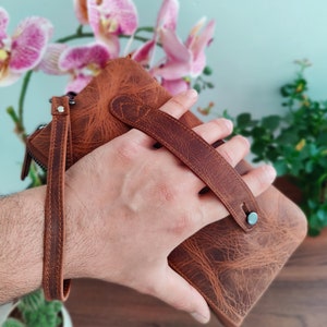 May include: A brown leather wallet with a strap and a metal clasp. The wallet is held in a hand.
