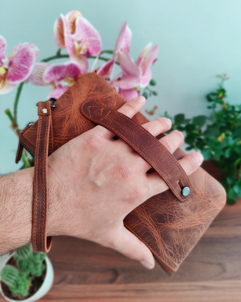 May include: A brown leather wallet with a strap and a metal clasp. The wallet is held in a hand.