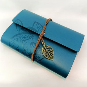 May include: Teal-colored journal with embossed leaf design. The journal has a flap closure and is secured with a brown leather cord. A gold-colored leaf charm hangs from the cord. The journal is a notebook.
