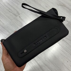 May include: Black leather clutch with a zipper closure and a top handle. The clutch has a small pocket on the front with the word "BASARAN" embossed in the leather.
