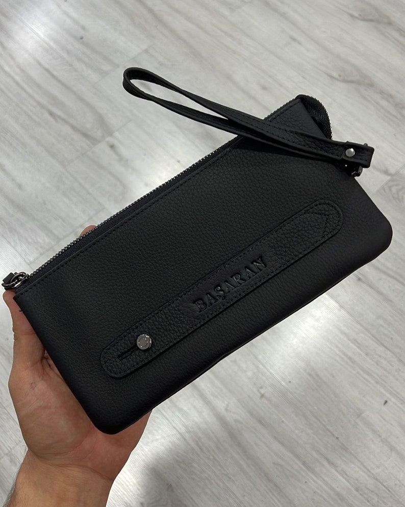 May include: Black leather clutch with a zipper closure and a top handle. The clutch has a small pocket on the front with the word "BASARAN" embossed in the leather.