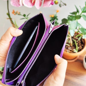 May include: A purple leather clutch with two zippered compartments. The clutch is open and a black phone is inside one of the compartments.