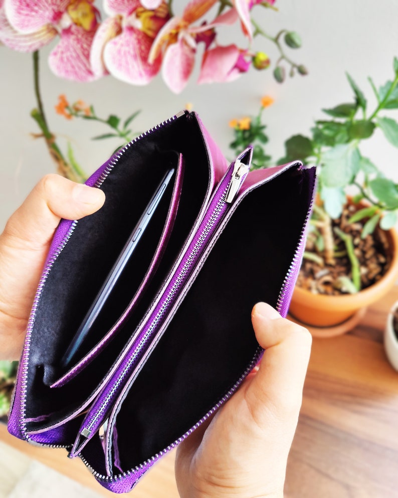May include: A purple leather clutch with two zippered compartments. The clutch is open and a black phone is inside one of the compartments.