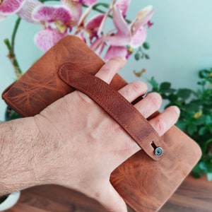 May include: A brown leather wallet with a strap handle. The wallet is held in a hand.