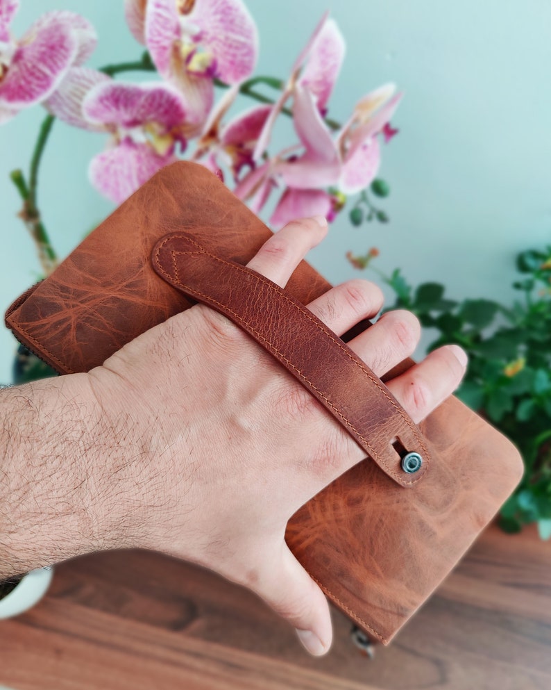May include: A brown leather wallet with a strap handle. The wallet is held in a hand.