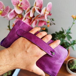May include: A purple leather wallet with a top handle and a zipper closure. The wallet is held in a hand.
