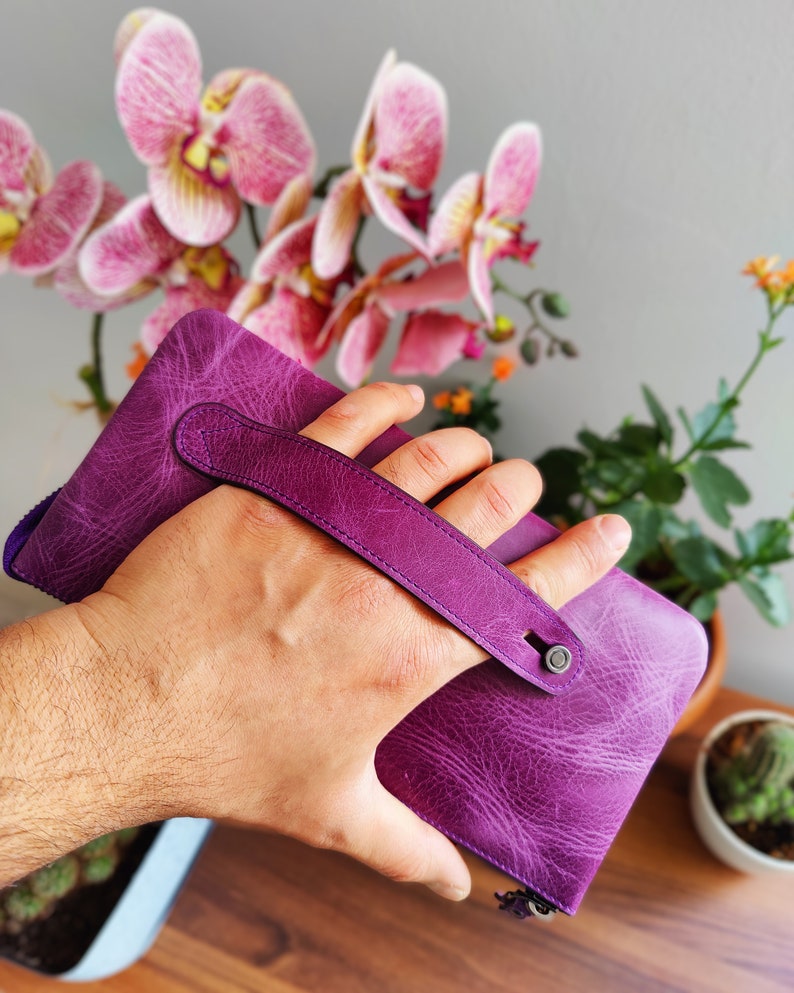 May include: A purple leather wallet with a top handle and a zipper closure. The wallet is held in a hand.