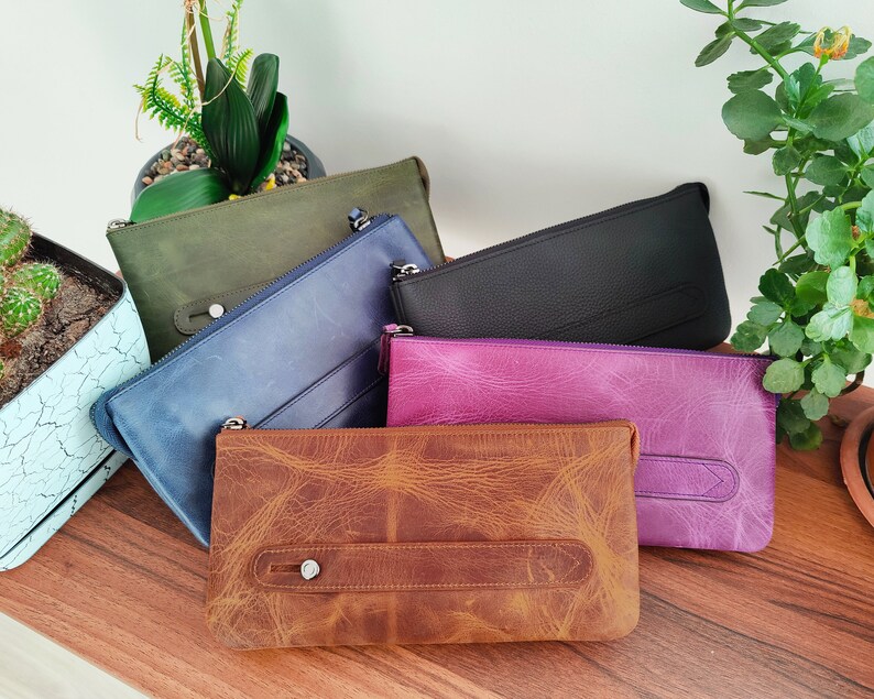 May include: Five leather pouches in different colors and textures. The pouches are arranged on a wooden surface. The pouches are all closed and have a zipper closure.