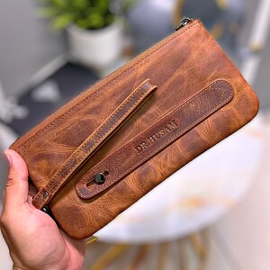 May include: A brown leather clutch with a zipper closure and a strap. The clutch has the name "DR. HUSAM" embossed on the front.