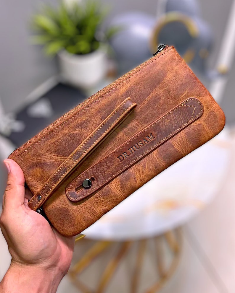 May include: A brown leather clutch with a zipper closure and a strap. The clutch has the name "DR. HUSAM" embossed on the front.