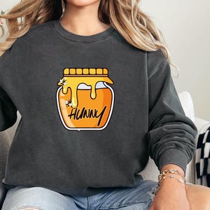 Puede incluir: Sudadera gris oscuro con un gráfico de un tarro de miel. El tarro está lleno de miel naranja y tiene la palabra "HUNNY" escrita. Dos abejas de dibujos animados zumban alrededor del tarro. La sudadera es de estilo casual.