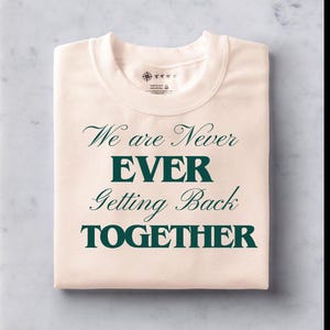 Könnte beinhalten: Ein gefaltetes, cremefarbenes Sweatshirt mit dem Text "We are Never EVER Getting Back TOGETHER" in dunkelgrüner Schrift. Das Shirt hat einen Rundhalsausschnitt und ein kleines Etikett in der Nähe des Kragens.