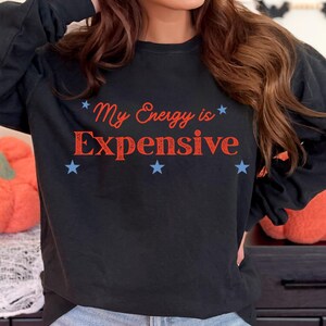 Puede incluir: Sudadera negra de manga larga con el texto "My Energy is Expensive" en rojo y azul, rodeado de estrellas azules. Una prenda de estilo casual.