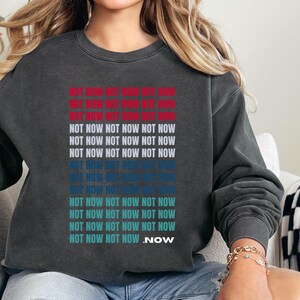 May include: Dark gray sweatshirt with the words "NOT NOW" repeated in red, white, and blue. The word "NOW" is at the end of the design. The sweatshirt is a casual style.