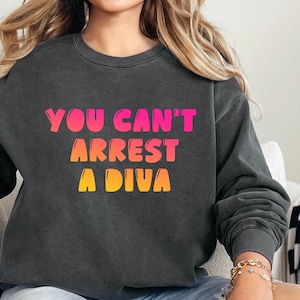Puede incluir: Sudadera gris oscuro con el texto "YOU CAN'T ARREST A DIVA" en degradado rosa, naranja y amarillo. Una prenda informal y cómoda para el uso diario.