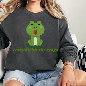 Peut inclure: Sweat-shirt gris foncé avec un motif de grenouille verte et le texte "I Stand with The Frogs". La grenouille est assise sur un nénuphar. Le sweat-shirt a des manches longues et un col rond.