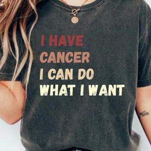 May include: Dark gray t-shirt with the text "I HAVE CANCER I CAN DO WHAT I WANT" in red, tan, and yellow lettering. The shirt has a vintage, worn look.