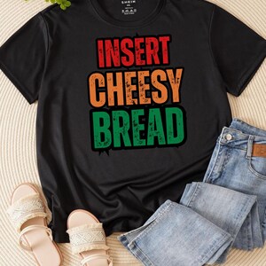 May include: Black t-shirt with the text "INSERT CHEESY BREAD" in red, orange, and green. The shirt is paired with light blue jeans and beige sandals. Sunglasses are also visible.