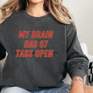 May include: Dark gray sweatshirt with the text "MY BRAIN HAS 67 TABS OPEN" in red letters with a yellow outline. The sweatshirt is worn with blue denim shorts. The person is wearing a gold bracelet.