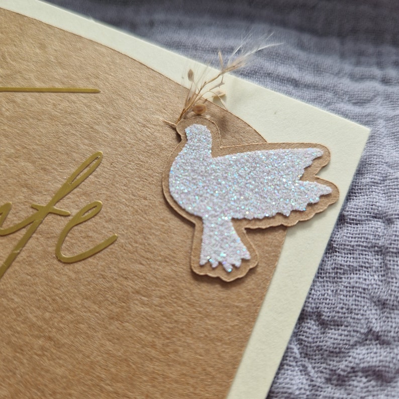 Op de afbeelding: Een bruine kaart met gouden letters die "Life" zeggen en een witte glitterduif met een takje gedroogde bloemen.