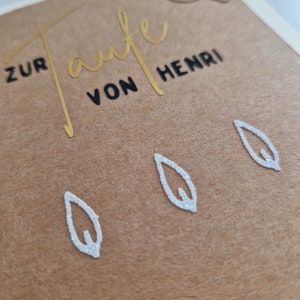 Op de afbeelding: Een bruine kaart met gouden letters die "Zur Taufe von Henri" zeggen. Er zijn drie witte bladeren met glitter en een witte duif met glitter op de kaart.