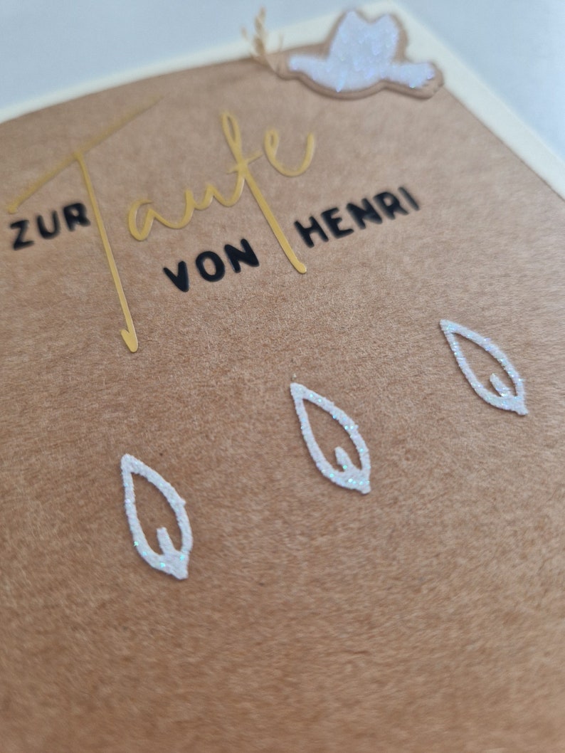 Op de afbeelding: Een bruine kaart met gouden letters die "Zur Taufe von Henri" zeggen. Er zijn drie witte bladeren met glitter en een witte duif met glitter op de kaart.