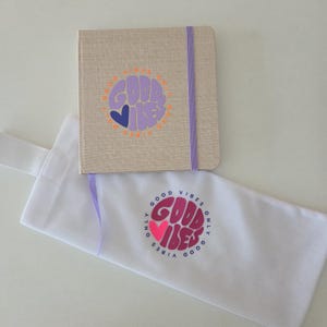 May include: A beige notebook with a purple elastic closure and the words "GOOD VIBES" in a circular design. A white tote bag with the same design is also present.