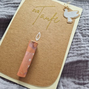 Op de afbeelding: Een bruine kaart met de woorden "zur Taufe" in gouden letters. Een witte duif met glitter is met touw aan de kaart bevestigd. Een kleine oranje kaars met witte sterren ligt op de kaart.