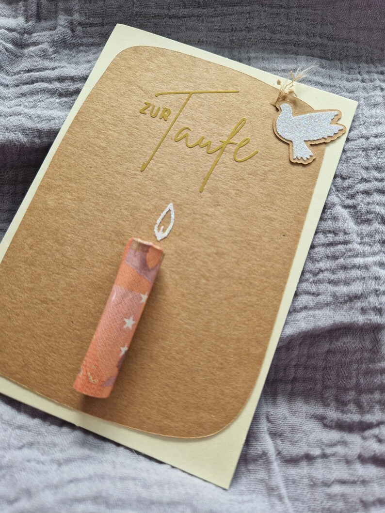 Op de afbeelding: Een bruine kaart met de woorden "zur Taufe" in gouden letters. Een witte duif met glitter is met touw aan de kaart bevestigd. Een kleine oranje kaars met witte sterren ligt op de kaart.
