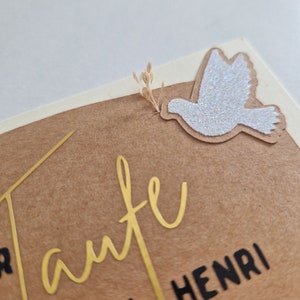 Op de afbeelding: Een bruine kaart met gouden letters die "Tante von Henri" zeggen en een witte glitterduif met een klein gedroogd bloemscherm.