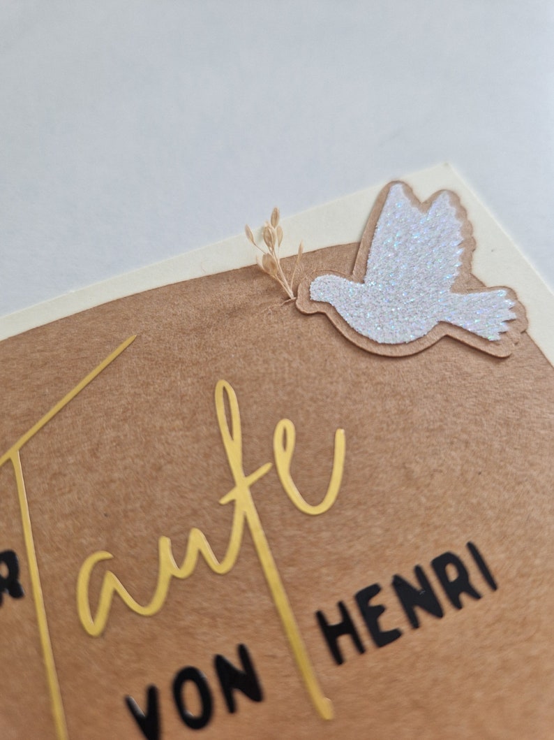 Op de afbeelding: Een bruine kaart met gouden letters die "Tante von Henri" zeggen en een witte glitterduif met een klein gedroogd bloemscherm.
