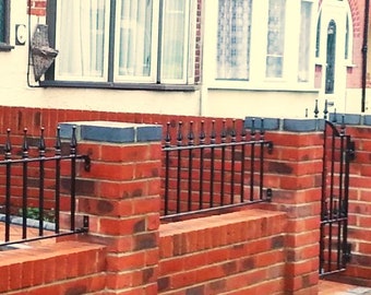May include: A red brick wall topped with a black wrought iron fence and gate. The wall has a dark grey cap. The fence features vertical bars and decorative finials. The gate is closed, providing a secure boundary.