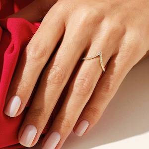 Könnte beinhalten: Ein goldener Ring mit einem V-förmigen Design, besetzt mit kleinen Diamanten. Der Ring wird am Mittelfinger einer Hand mit hellrosa Nagellack getragen.