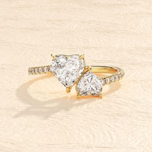 May include: A gold ring featuring two heart-shaped diamonds. The larger diamond is set in the centre of the ring, with the smaller diamond set on the side. The band is adorned with smaller diamonds.
