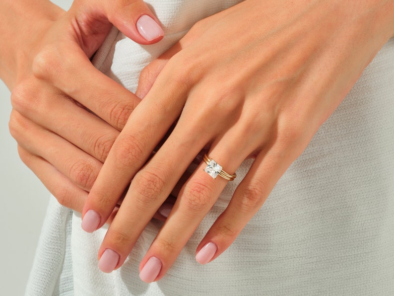 May include: A close-up of a hand wearing a gold ring with a large, square-shaped diamond. The ring is set with smaller diamonds on the band.