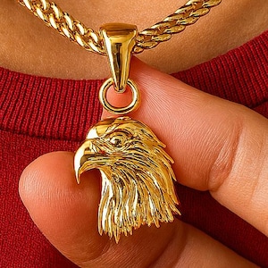 May include: Gold-colored eagle pendant necklace. The pendant is a detailed rendering of an eagle's head. The necklace chain is a thick, textured gold. The background is a red shirt.