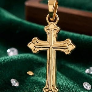 Puede incluir: Un colgante dorado con forma de cruz, con detalles ornamentados. La cruz tiene un lazo en la parte superior para una cadena. Se exhibe sobre una superficie de terciopelo verde oscuro, con peque&ntilde;os elementos decorativos.
