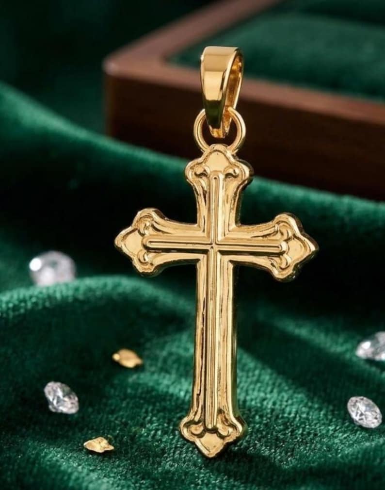 Puede incluir: Un colgante dorado con forma de cruz, con detalles ornamentados. La cruz tiene un lazo en la parte superior para una cadena. Se exhibe sobre una superficie de terciopelo verde oscuro, con peque&ntilde;os elementos decorativos.