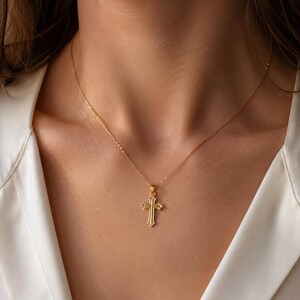 Puede incluir: Un collar con colgante de cruz de color dorado. La cruz tiene un dise&ntilde;o detallado con una superficie texturizada y est&aacute; suspendida de una delicada cadena. El collar se lleva sobre una camisa blanca.