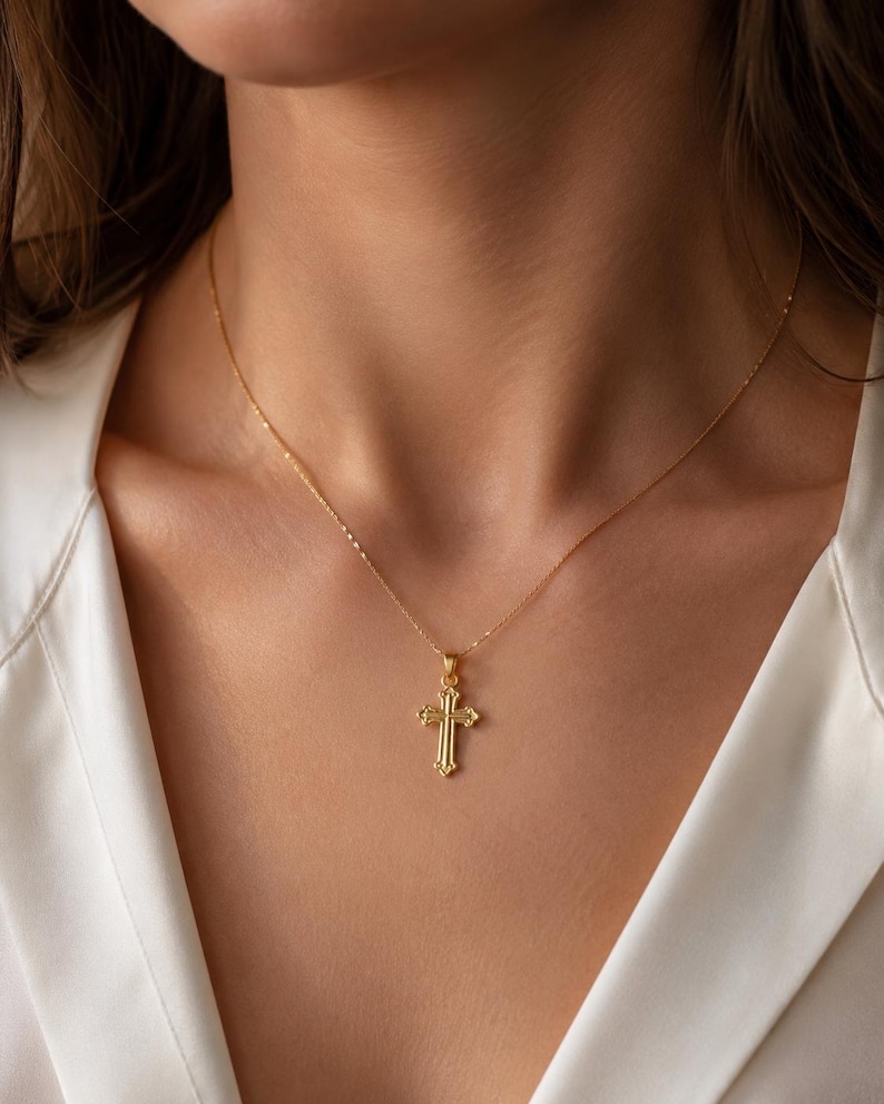 Puede incluir: Un collar con colgante de cruz de color dorado. La cruz tiene un dise&ntilde;o detallado con una superficie texturizada y est&aacute; suspendida de una delicada cadena. El collar se lleva sobre una camisa blanca.