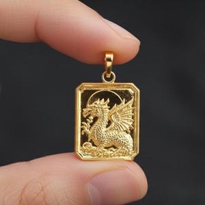Puede incluir: Colgante dorado con forma rectangular y diseño de dragón. El dragón está detallado con alas y escamas. El colgante se sostiene entre los dedos sobre un fondo oscuro.