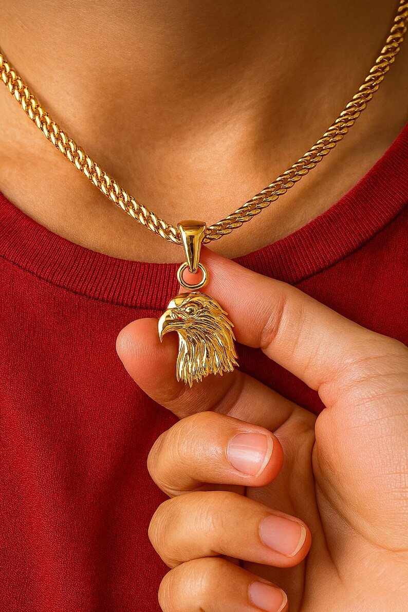 May include: Gold-colored eagle pendant necklace. The pendant is a detailed rendering of an eagle's head. The necklace chain is a thick, textured gold. The background is a red shirt.