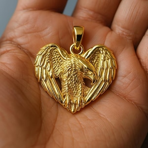 May include: A gold-coloured heart-shaped pendant, depicting an eagle with spread wings. The eagle's head is central, with wings extending outwards. The pendant is held in a hand, showing the intricate details.