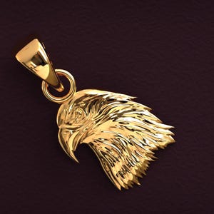 May include: A gold-colored pendant shaped like an eagle's head. The pendant features detailed feather textures and a loop for a chain. The eagle's beak is slightly open, and the overall design is highly polished.