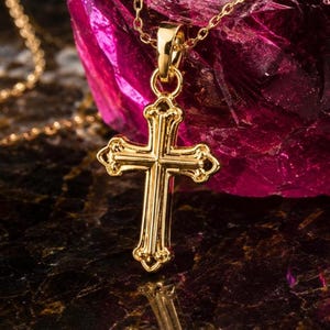 Puede incluir: Un colgante de cruz dorada con un dise&ntilde;o ornamentado cuelga de una delicada cadena. La cruz tiene bordes decorativos y un acabado pulido. El fondo presenta una superficie oscura y reflectante y un objeto magenta grande y texturizado.