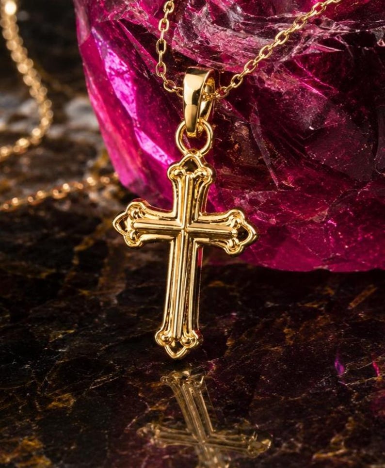 Puede incluir: Un colgante de cruz dorada con un dise&ntilde;o ornamentado cuelga de una delicada cadena. La cruz tiene bordes decorativos y un acabado pulido. El fondo presenta una superficie oscura y reflectante y un objeto magenta grande y texturizado.