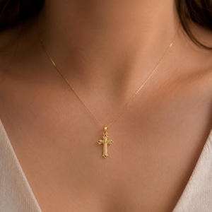 Puede incluir: Un collar con colgante dorado con un dise&ntilde;o de cruz detallado. La cruz tiene detalles ornamentados y cuelga de una delicada cadena dorada. El collar se muestra sobre un fondo neutro.