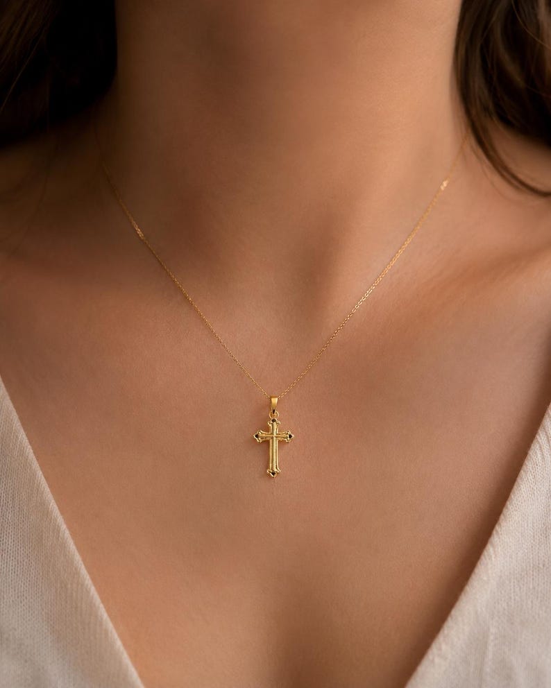 Puede incluir: Un collar con colgante dorado con un dise&ntilde;o de cruz detallado. La cruz tiene detalles ornamentados y cuelga de una delicada cadena dorada. El collar se muestra sobre un fondo neutro.