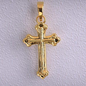 Puede incluir: Un colgante dorado con forma de cruz. La cruz tiene detalles ornamentados y un lazo en la parte superior para una cadena. El colgante se muestra sobre un fondo texturizado blanquecino.